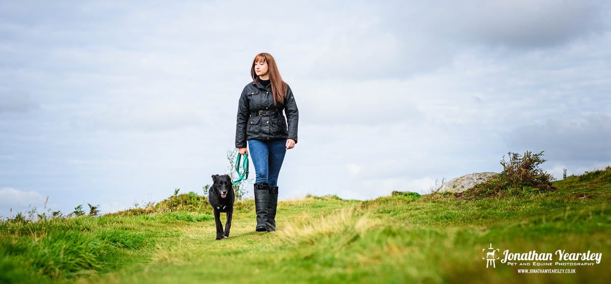 A woman with long brown hair, wearing a black jacket and boots, walks a black dog on a grassy hill under a cloudy sky. She holds a blue leash as the green foreground features scattered bushes.